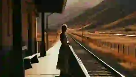 A woman standing at the edge of a train station platform, looking out at the tracks and the distant hills in the sunset.