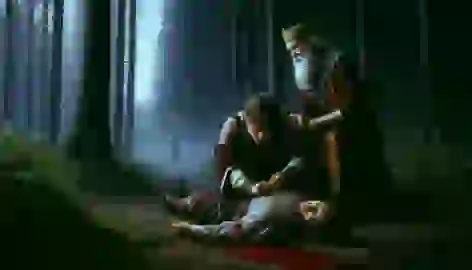 The king kneels beside a wounded man, bandaging his side while the hermit stands watching in concern.